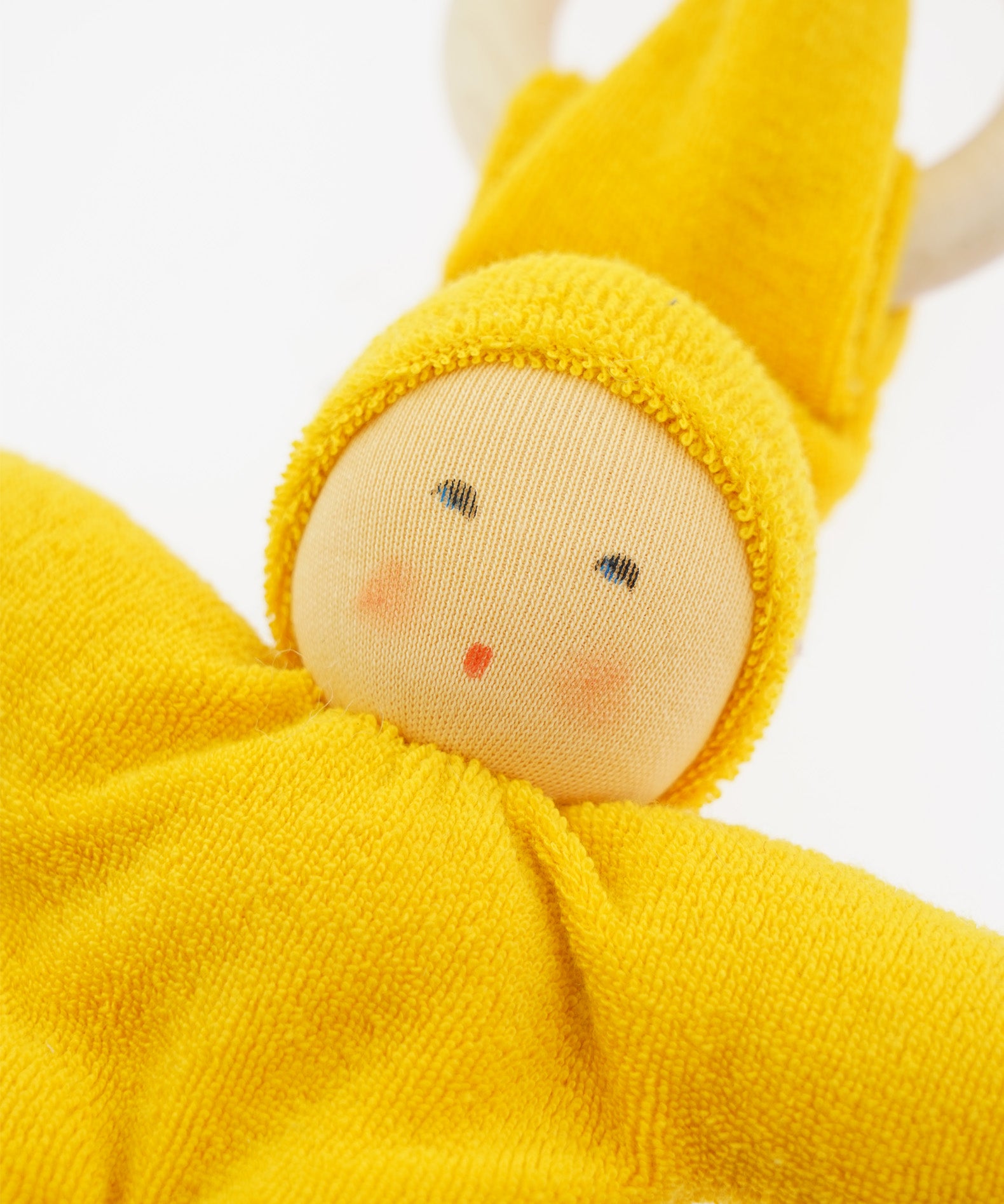 Close-up of the yellow Nanchen Waldorf Pimpel Doll. The doll has a hand-painted face with rosy cheeks and a small red mouth, surrounded by a bright yellow cotton terry hood and body. A natural wooden teething ring is seen in the background.