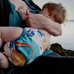 A breastfeeding baby wearing The Close Pop-In x Babipur exclusive reusable nappy wrap, in a Babipur World Print