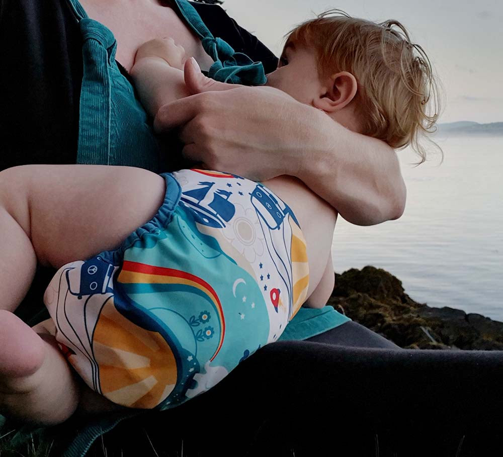 A breastfeeding baby wearing The Close Pop-In x Babipur exclusive reusable nappy wrap, in a Babipur World Print