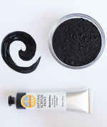 A top down view of the Natural earth paint in black ochre next to a bowl of black pigment and a paint streak showing the consistency. 