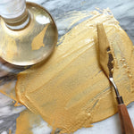 Natural Earth Paint palette knife with wooden handle spreading gold glittery paint on a stone surface. 