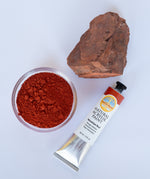 A top down view of the Natural earth paint tube in venetian red next to a rock covered in matching paint pigment and a small glass bowl of pigment