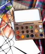The  Natural Earth Paint small wooden watercolour palette on top of a canvas with a abstract painting showing different coloured paint in the well and little magnetic pieces on each corner to open and close 