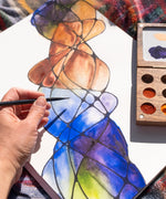 A close up of the Natural Earth Paint small wooden watercolour palette on top of a canvas with an adult hand holding a paint brush showing the scale of the palette. 