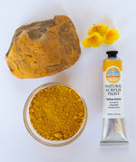 A top down view of the Natural earth paint tube in yellow ochre next to a rock covered in matching paint pigment and little yellow flowers