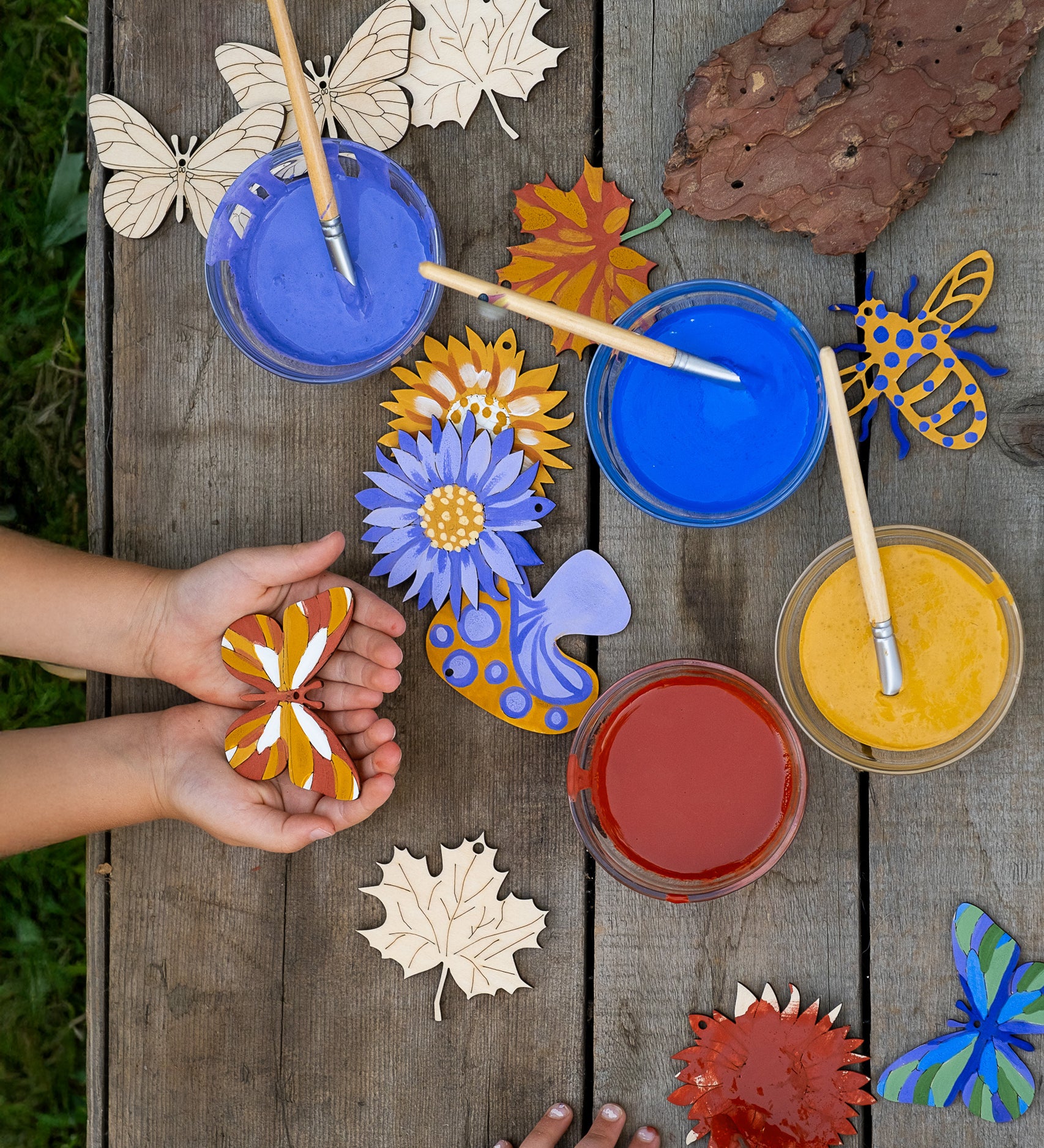 An overhead view of children using the Natural Earth Paint nature craft kit. they can be seen painting and holding their painted wooden decorations