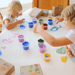 A top down view of 2 kids being creative using the Neogrun crystal finger paints on a white table cloth.