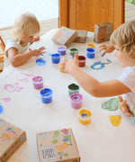 A top down view of 2 kids being creative using the Neogrun crystal finger paints on a white table cloth.