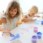 A top down view of 2 kids being creative and painting a picture on a white table cloth using the Neogrun crystal finger paints