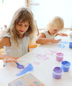 A top down view of 2 kids being creative and painting a picture on a white table cloth using the Neogrun crystal finger paints