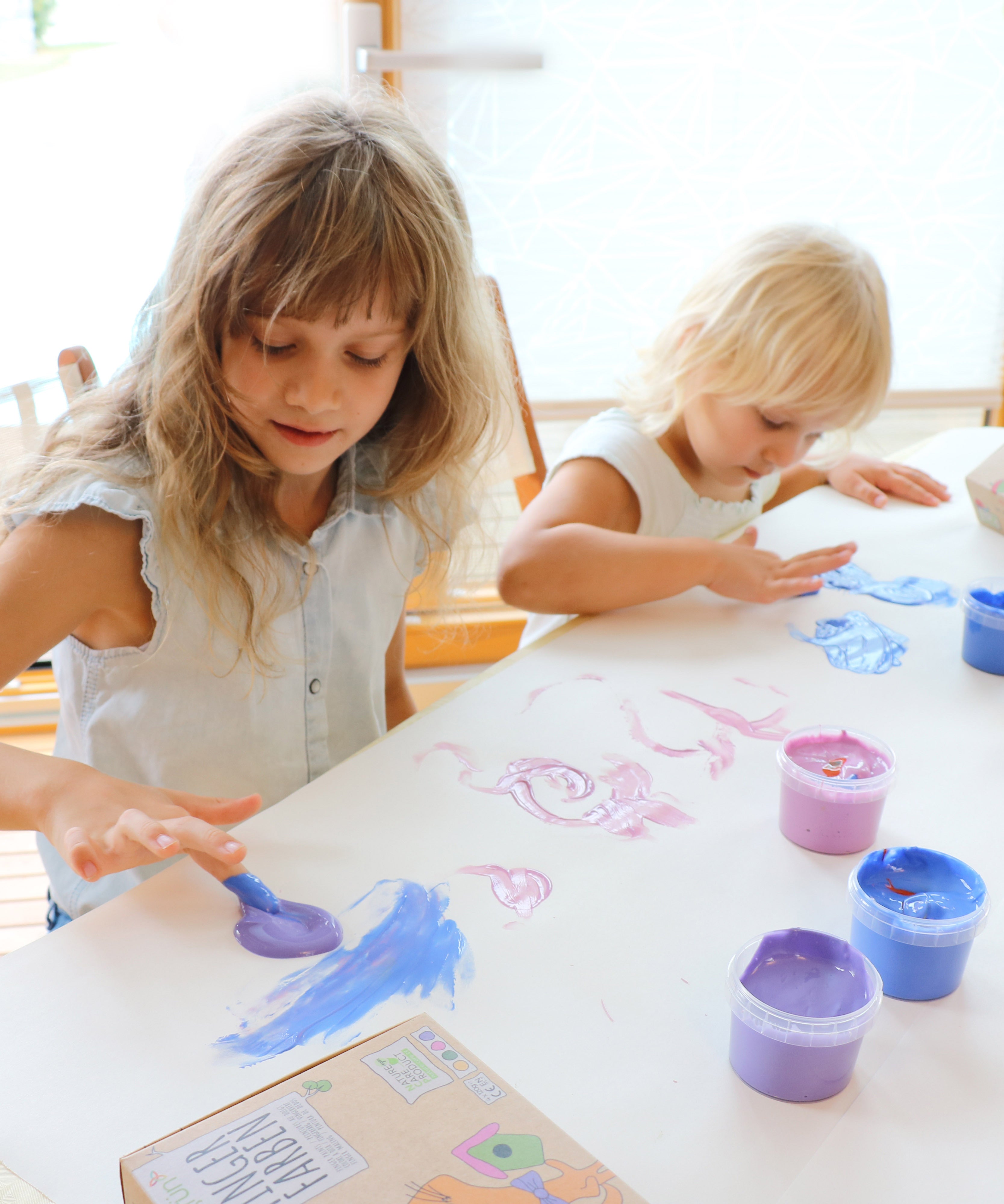 A top down view of 2 kids being creative and painting a picture on a white table cloth using the Neogrun crystal finger paints