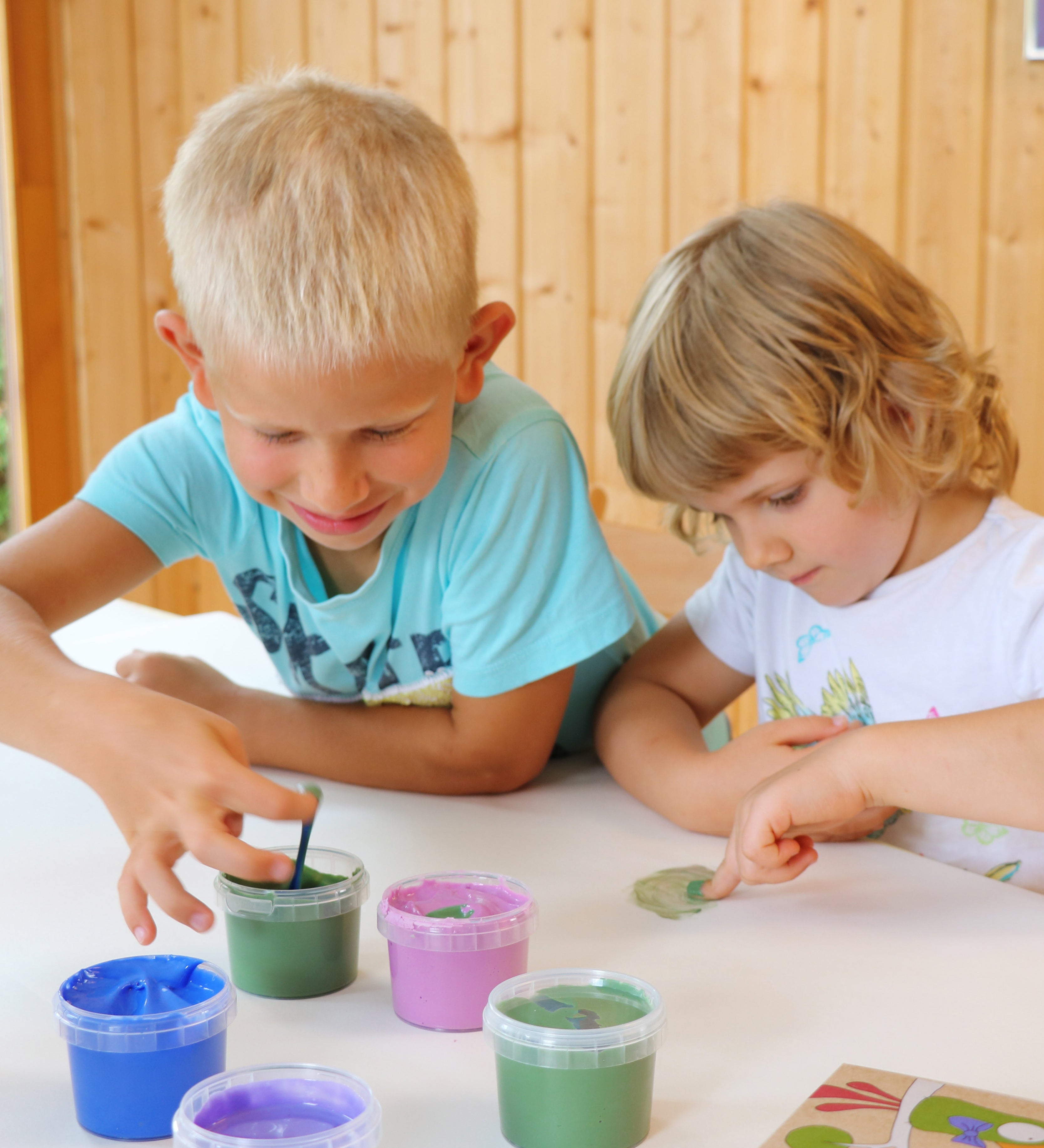 Two kids with their fingers in the Neogrun finger paint in the crystal colour set. showing 2 kids being creative. Features a pink, green blue and purple coloured paint. 