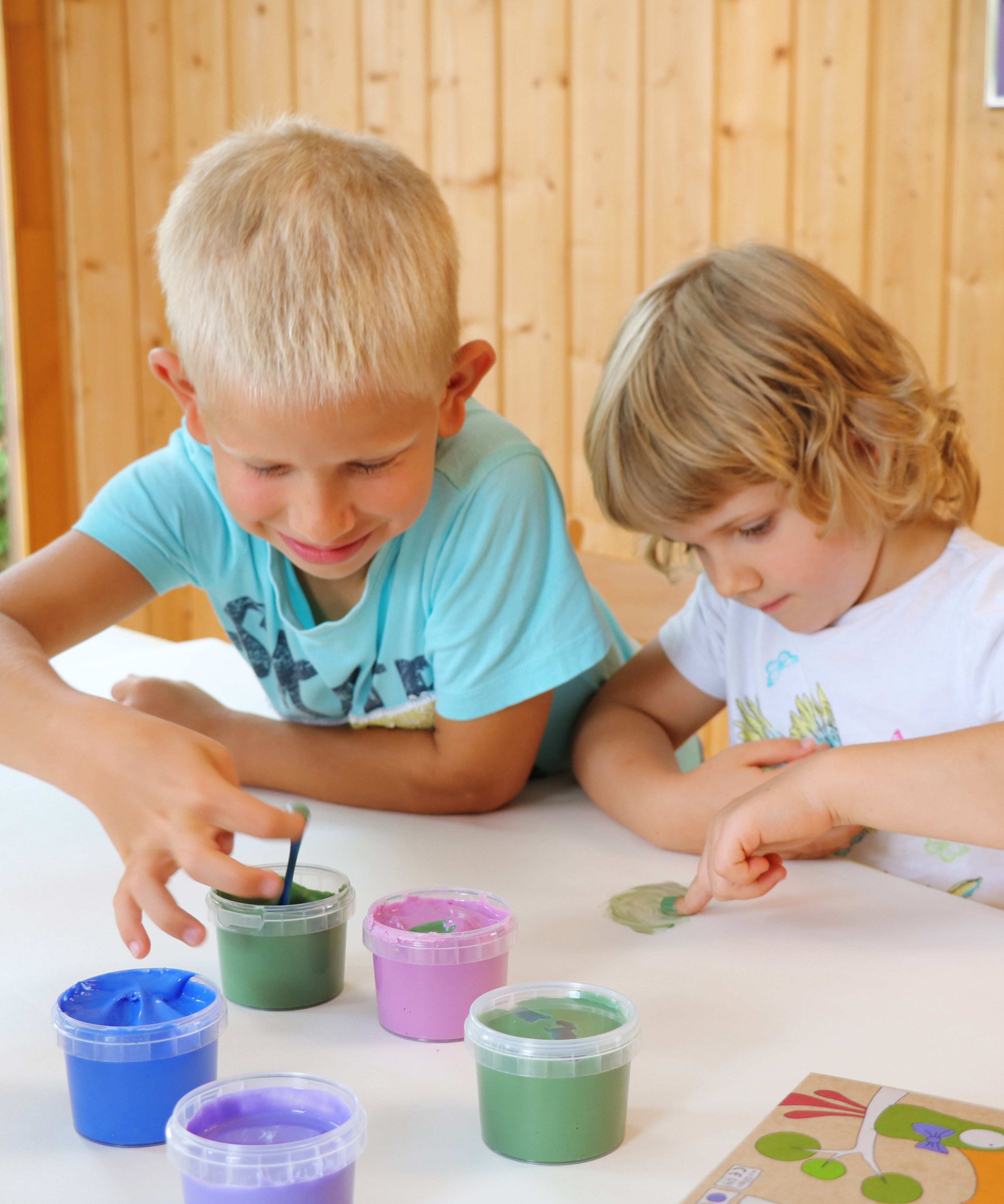 Two kids with their fingers in the Neogrun finger paint in the crystal colour set. showing 2 kids being creative. Features a pink, green blue and purple coloured paint. 