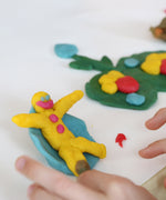 A close up of a child's hand being creative and moulded the Neogrun organic play dough into a little figure on a blue seat. 