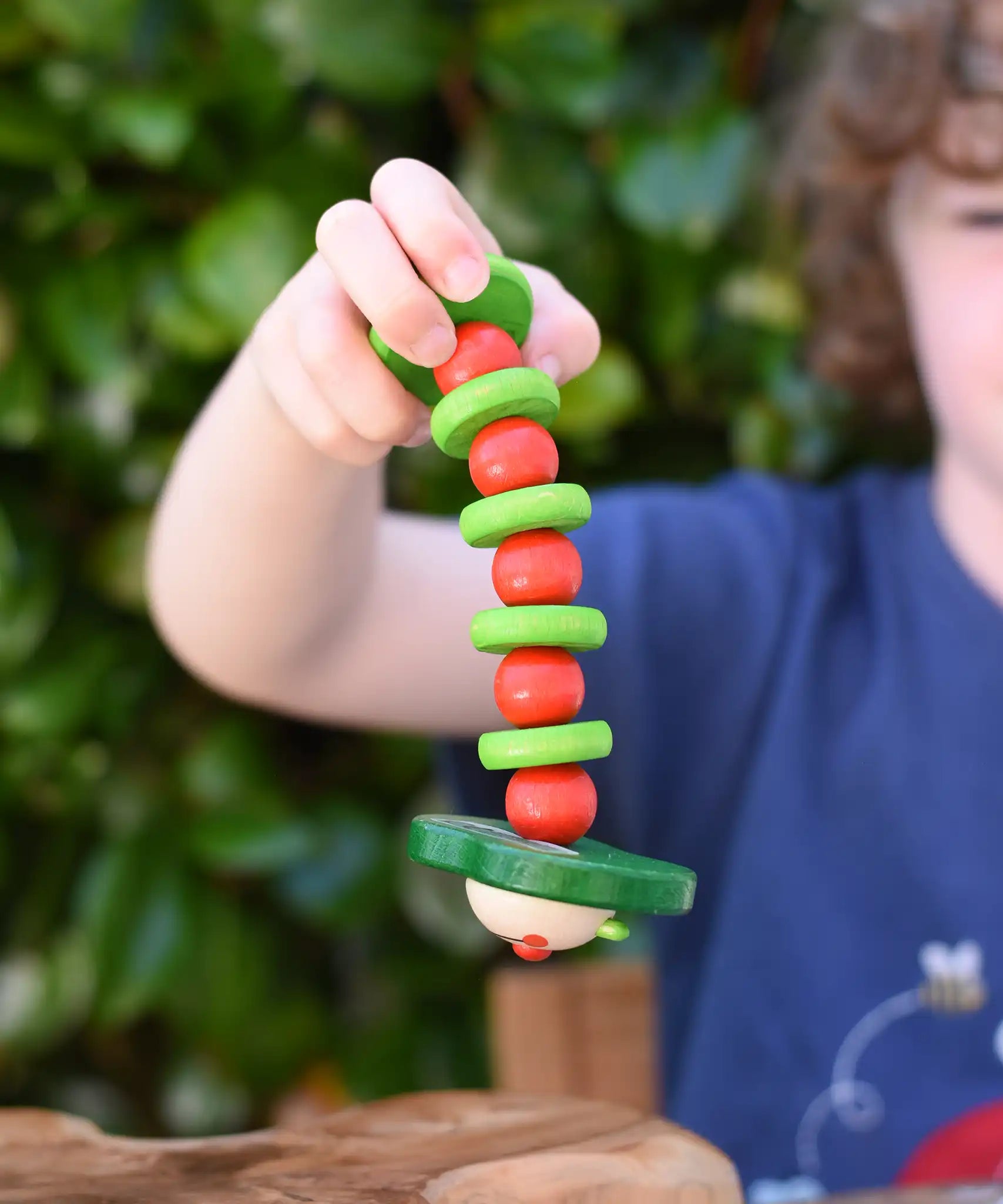 Nic apple worm baby wooden grasping toy in a child's hand. Smiling worm on a flexible rope lined with chunky red and green beads.