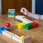 The Nic Cubio wooden starter ball track set shown set up on a wooden table with the outlet piece with ringing bell added to the end. The back on the outlet can be seen closest to the camera with a yellow ball at the end and a red coloured ball on the track.