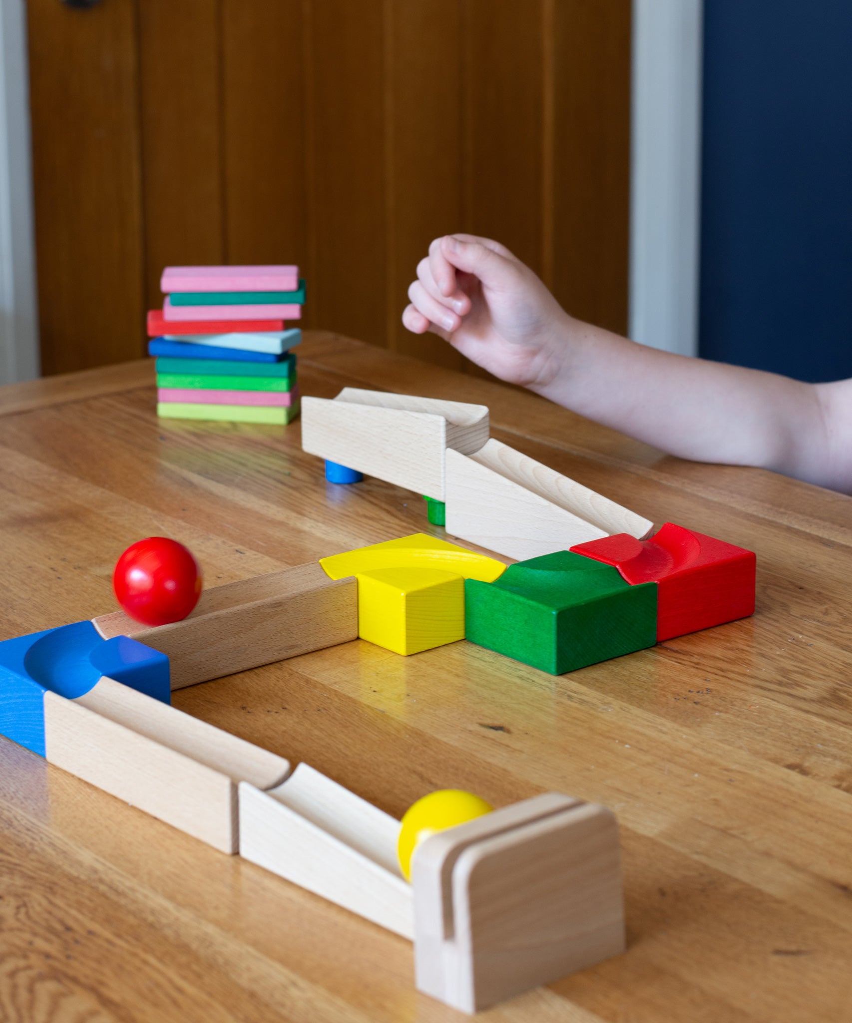 The Nic Cubio wooden starter ball track set shown set up on a wooden table with the outlet piece with ringing bell added to the end. The back on the outlet can be seen closest to the camera with a yellow ball at the end and a red coloured ball on the track.