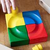 Four coloured corner pieces from the Nic Cubio Wooden ball run set placed together to create a simple circular shaped ball run track. A child's hand can be seen placing the yellow ball on the track. Other pieces from the track can be seen at the sides. 