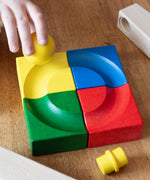 Four coloured corner pieces from the Nic Cubio Wooden ball run set placed together to create a simple circular shaped ball run track. A child's hand can be seen placing the yellow ball on the track. Other pieces from the track can be seen at the sides. 
