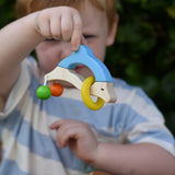 Nic dolphin wooden baby grasping toy n a child's hand. A blue and white dolphin with a green and orange bead attached to its tails and a yellow ring on the body.