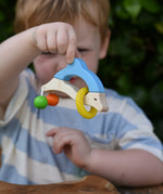 Nic dolphin wooden baby grasping toy n a child's hand. A blue and white dolphin with a green and orange bead attached to its tails and a yellow ring on the body.