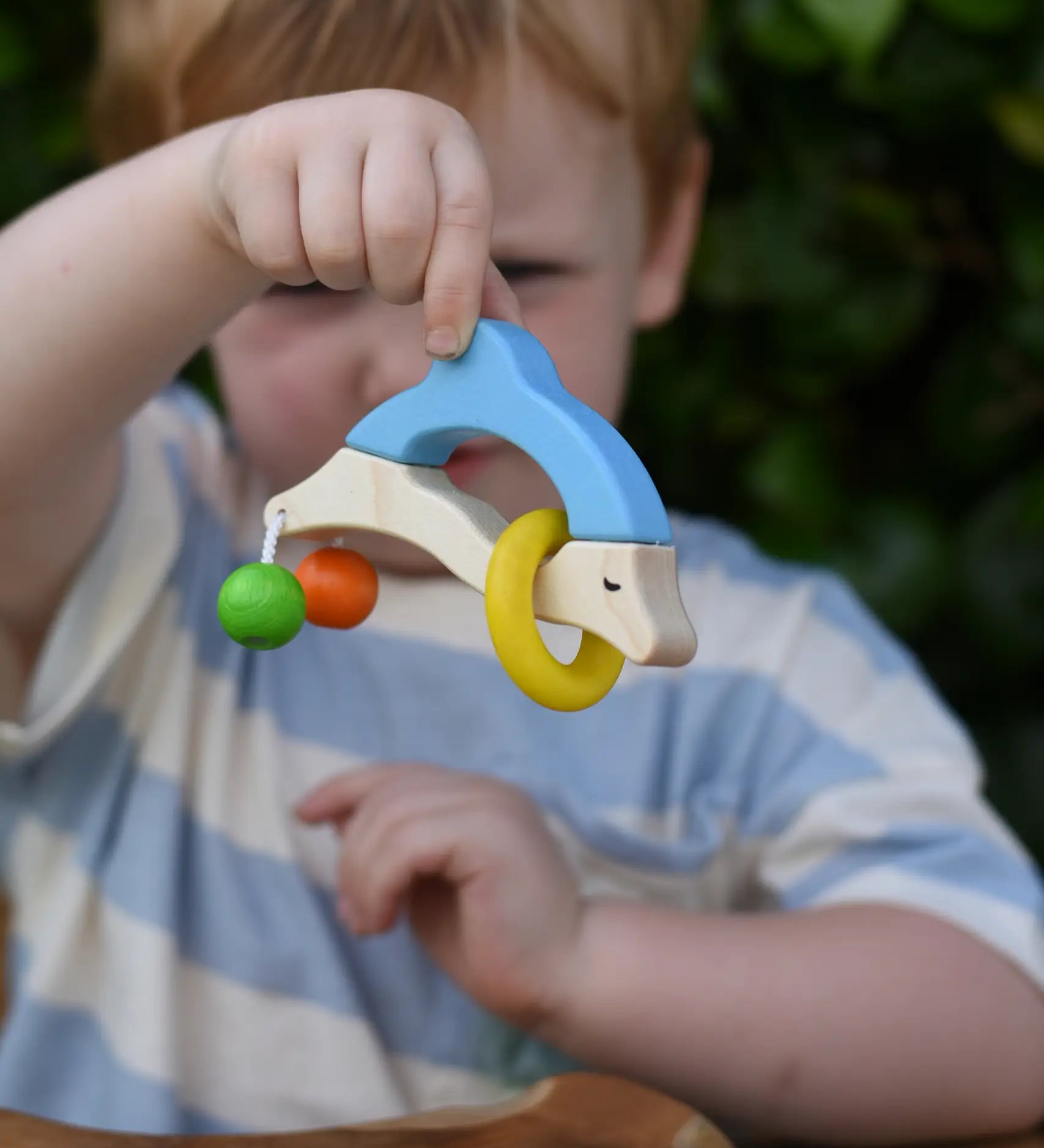 Nic dolphin wooden baby grasping toy n a child's hand. A blue and white dolphin with a green and orange bead attached to its tails and a yellow ring on the body.