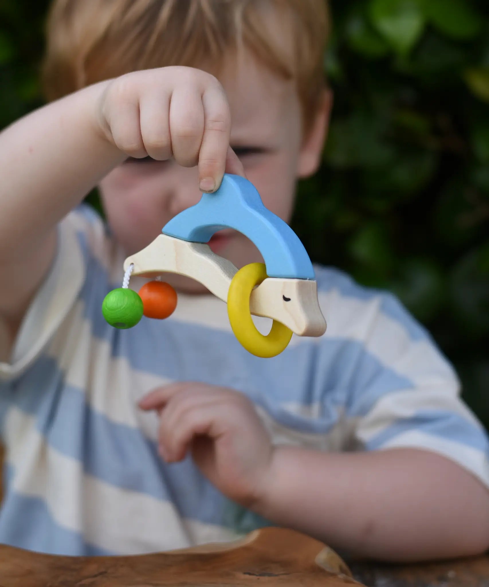 Nic dolphin wooden baby grasping toy n a child's hand. A blue and white dolphin with a green and orange bead attached to its tails and a yellow ring on the body.