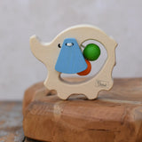 Nic elephant grasper wooden baby toy placed on a wooden surface. A light wood elephant body with blue ears and two green and orange beads inside. 