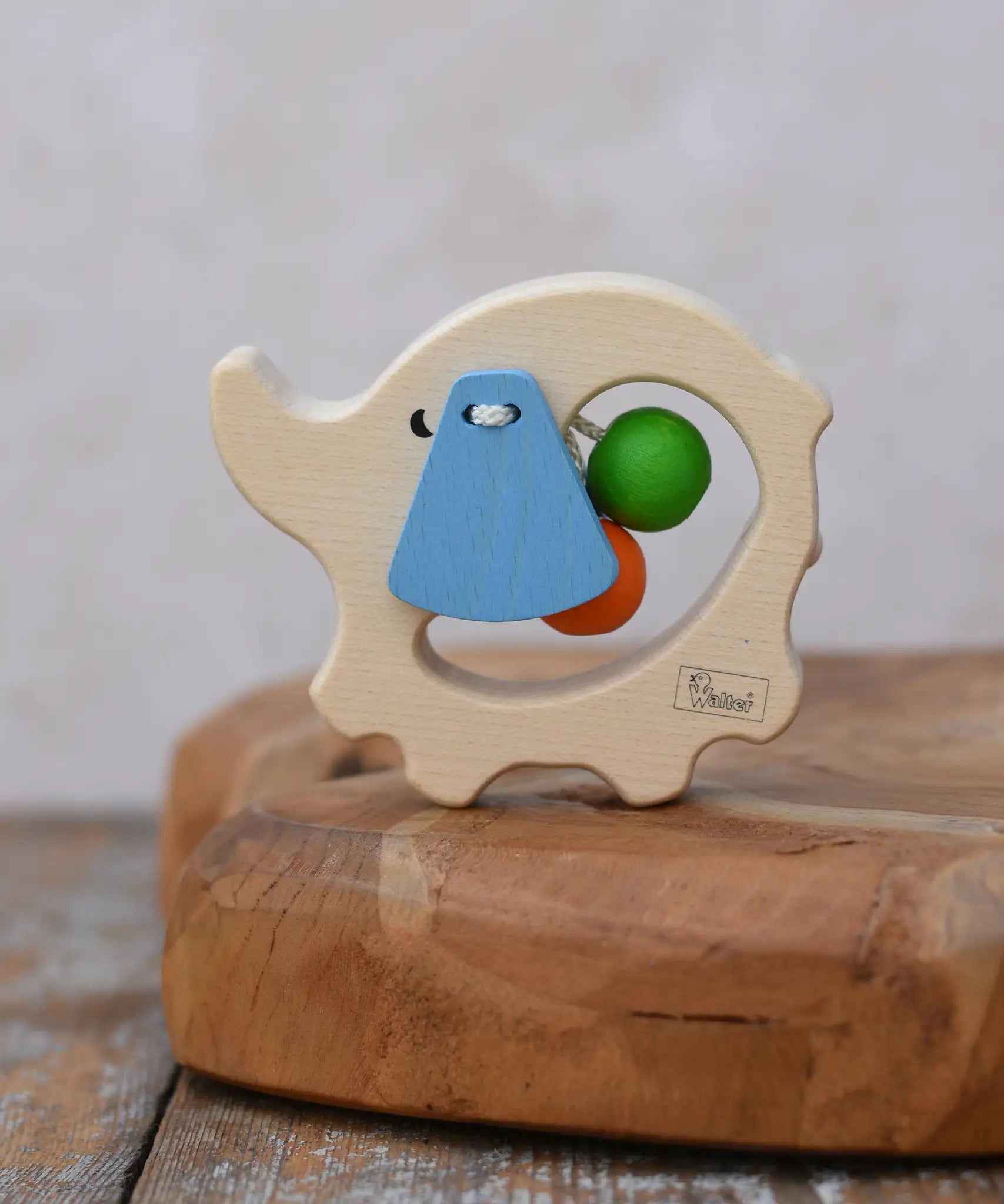 Nic elephant grasper wooden baby toy placed on a wooden surface. A light wood elephant body with blue ears and two green and orange beads inside. 
