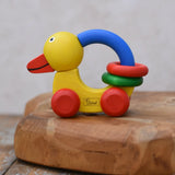 Nic Toys wooden grip duck in yellow with red wheels placed on a wooden dish. Features a blue handle for grip with red and green rings for play.  