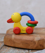 Nic Toys wooden grip duck in yellow with red wheels placed on a wooden dish. Features a blue handle for grip with red and green rings for play.  