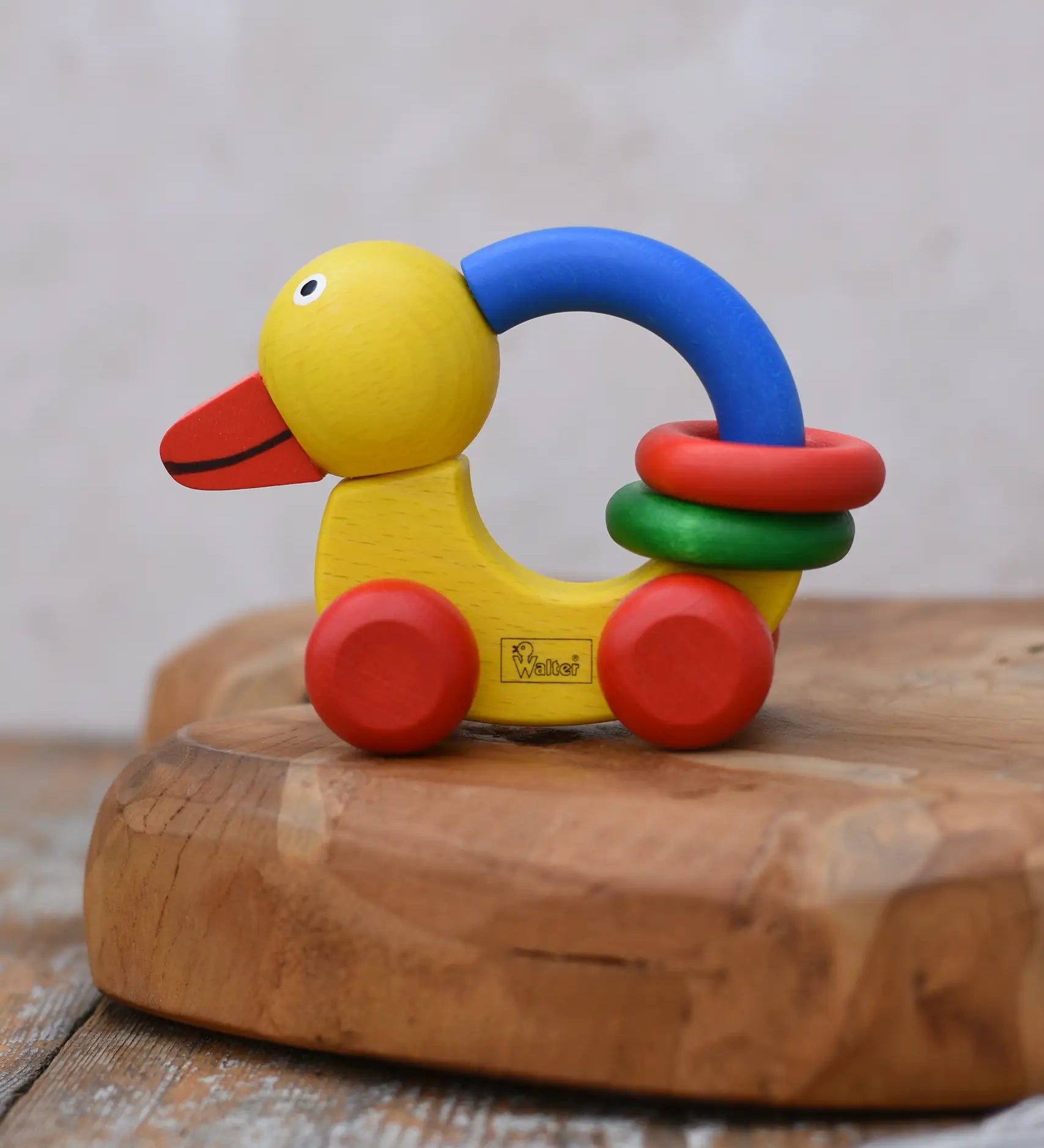Nic Toys wooden grip duck in yellow with red wheels placed on a wooden dish. Features a blue handle for grip with red and green rings for play.  