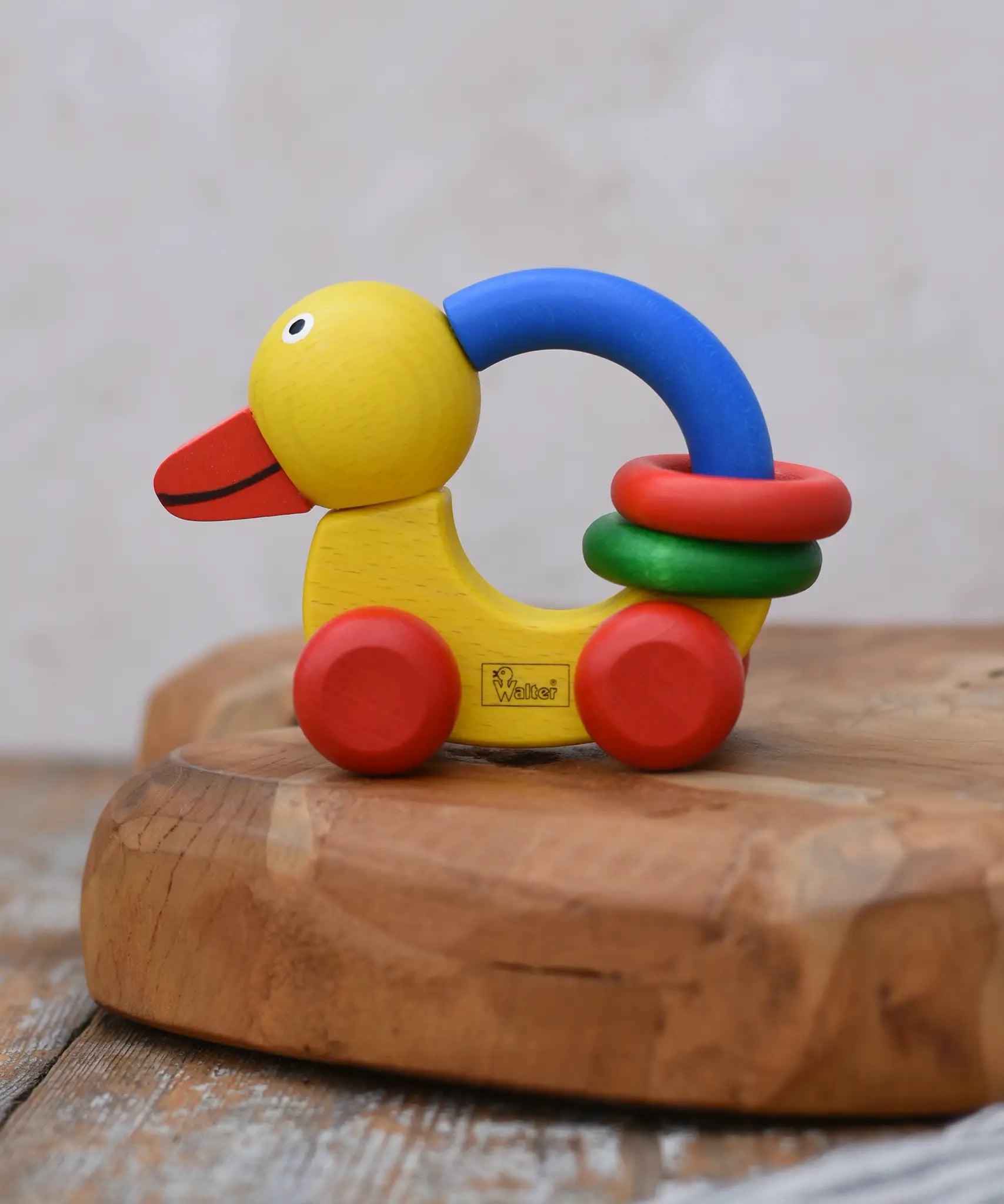 Nic Toys wooden grip duck in yellow with red wheels placed on a wooden dish. Features a blue handle for grip with red and green rings for play.  