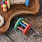 Nic organic jingle bell wooden roller baby toy placed on a wooden surface next to a wooden dish filled with colourful baby toys. 