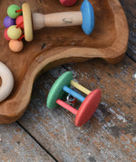 Nic organic jingle bell wooden roller baby toy placed on a wooden surface next to a wooden dish filled with colourful baby toys. 