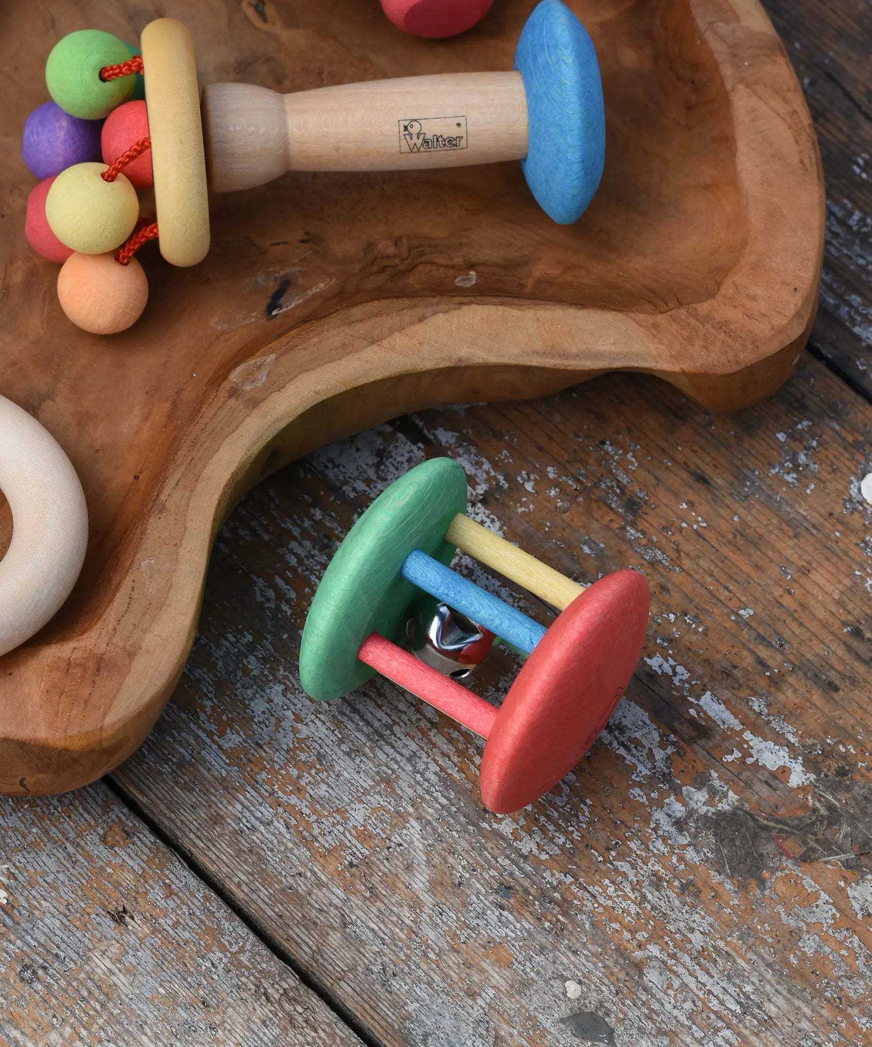 Nic organic jingle bell wooden roller baby toy placed on a wooden surface next to a wooden dish filled with colourful baby toys. 
