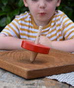Nic Toys wooden red spinning top placed on a Mader oak spinning board. Features a wooden handle for handling the spinner top with a blue cord for spinning the top. 