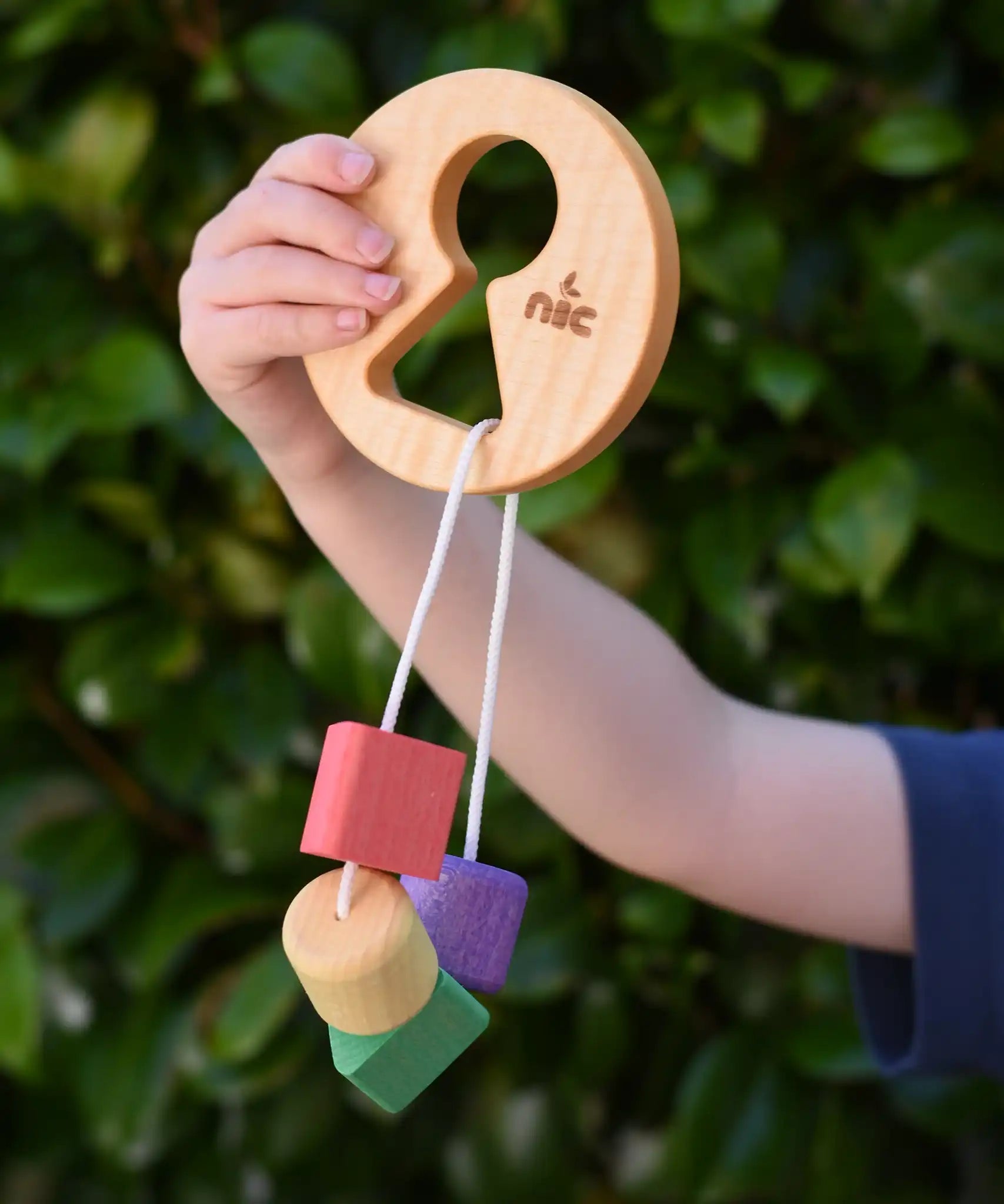 Child holding up the Nic Toys wooden threaded shape sorter. Wooden disk with a keyhole shape cut out in the middle with a piece of thread feeding through different coloured blocks in different coloured shapes.