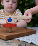 Nic Toys wooden red spinning top on an oak Mace spinning plate. Features a wooden handle for handling the spinner top with a blue cord for spinning the top. 