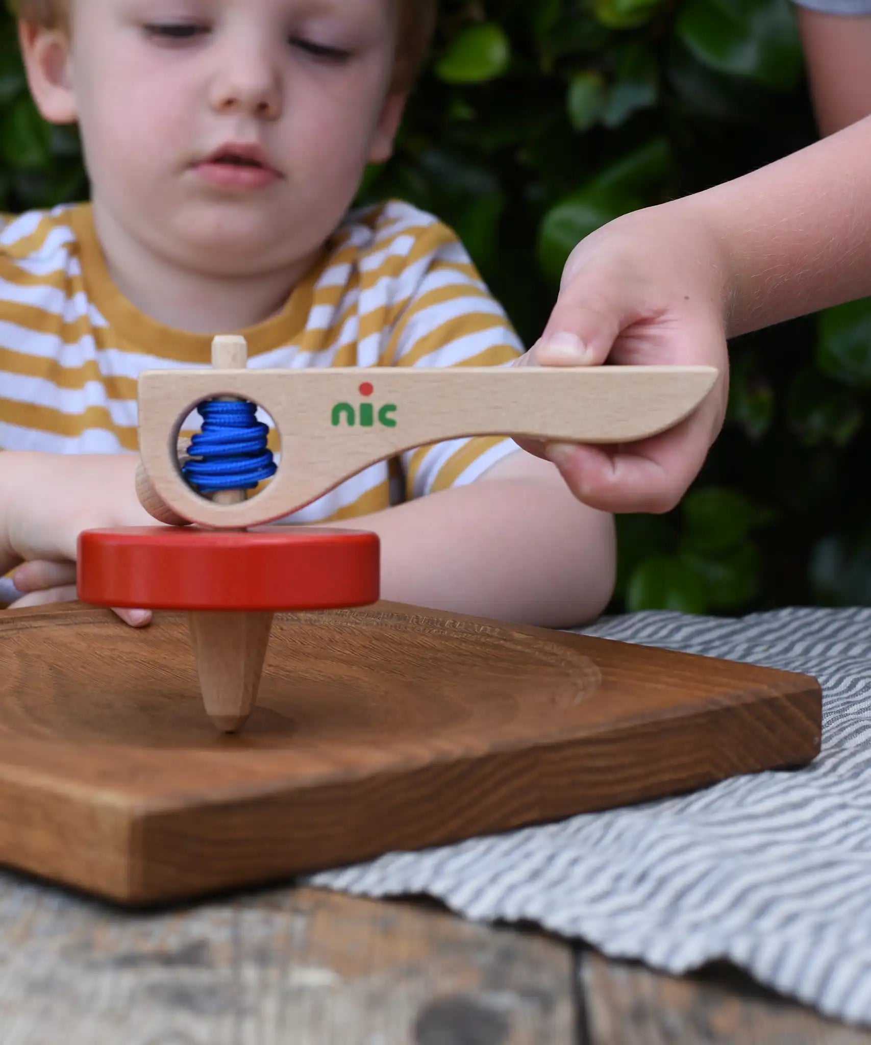 Nic Toys wooden red spinning top on an oak Mace spinning plate. Features a wooden handle for handling the spinner top with a blue cord for spinning the top. 