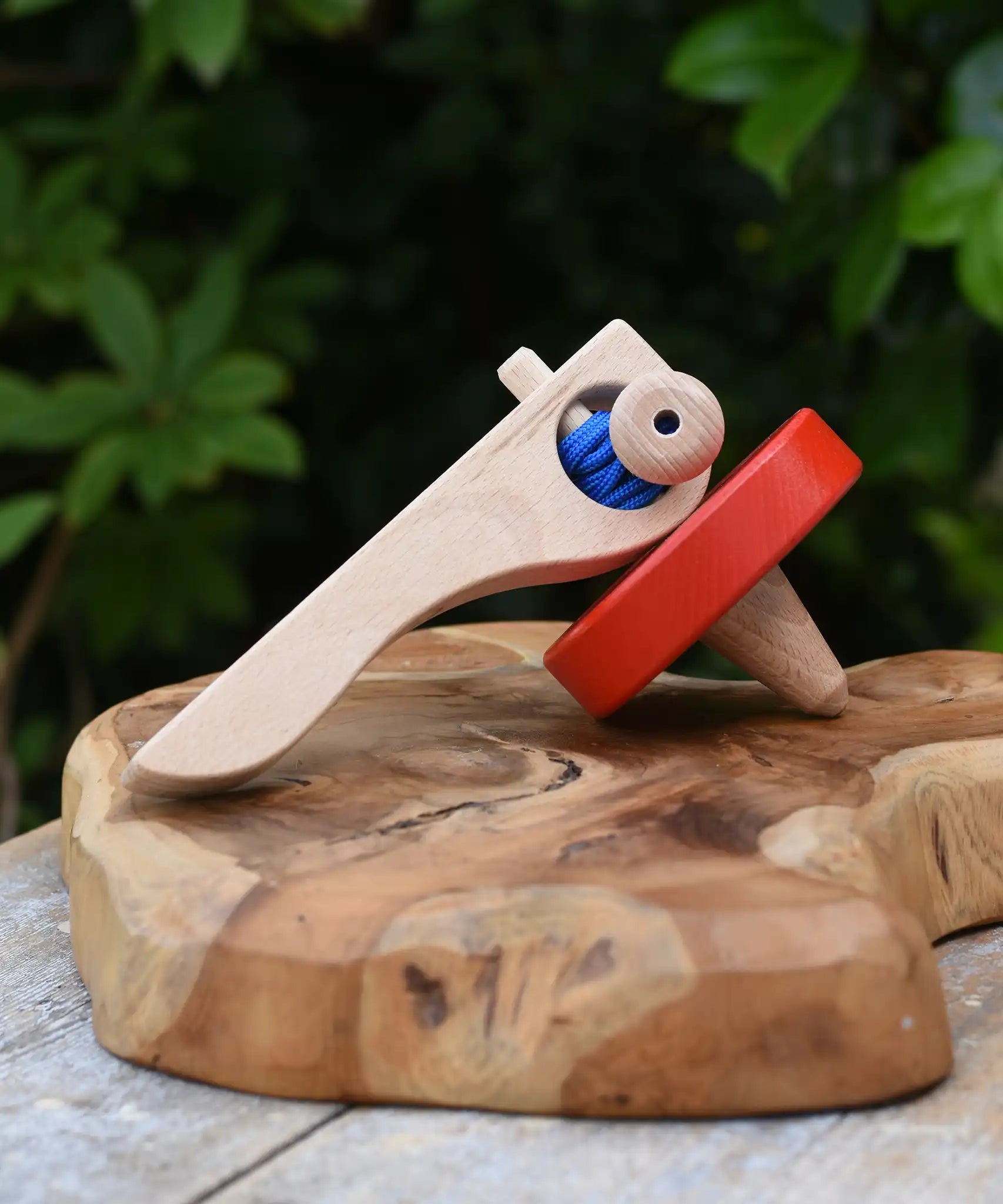 Nic Toys wooden red spinning top placed on a wooden surface outdoors. Features a wooden handle for handling the spinner top with a blue cord for spinning the top. 