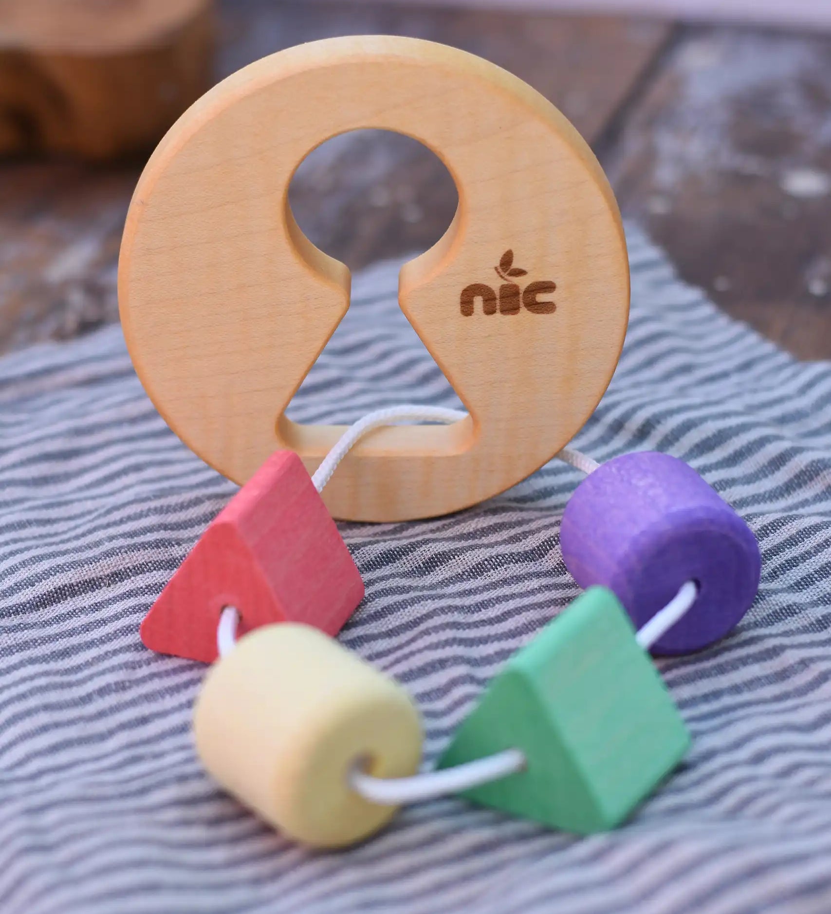 Nic Toys wooden threaded shape sorter placed on a cloth on a wooden surface. Wooden disk with a keyhole shape cut out in the middle with a piece of thread feeding through different coloured blocks in different coloured shapes.