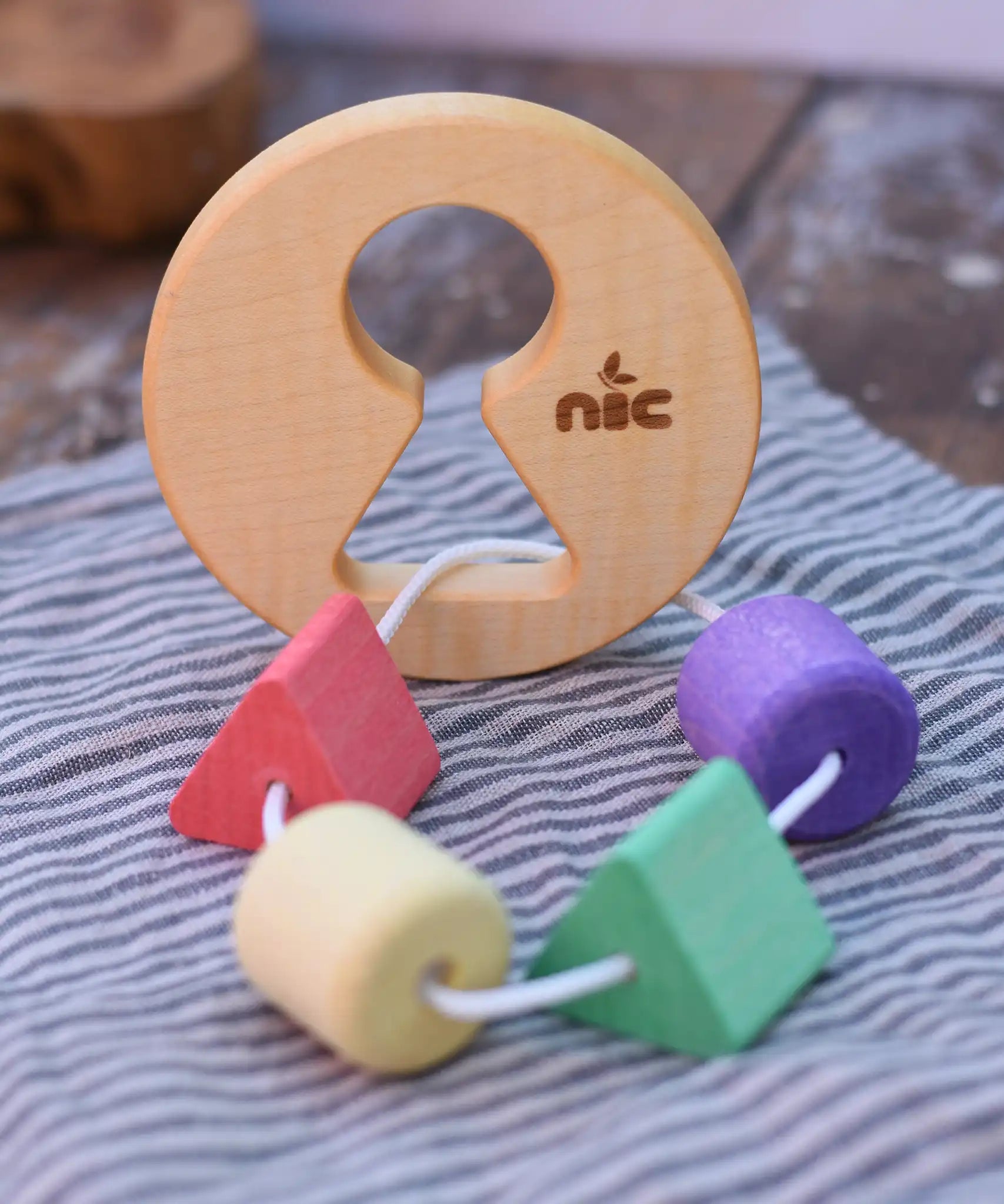 Nic Toys wooden threaded shape sorter placed on a cloth on a wooden surface. Wooden disk with a keyhole shape cut out in the middle with a piece of thread feeding through different coloured blocks in different coloured shapes.