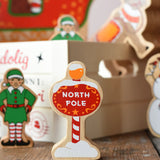 Lanka Kade red north pole sign wooden toy figure placed on a wooden surface in the Babipur playroom with other festive figures in teh background.