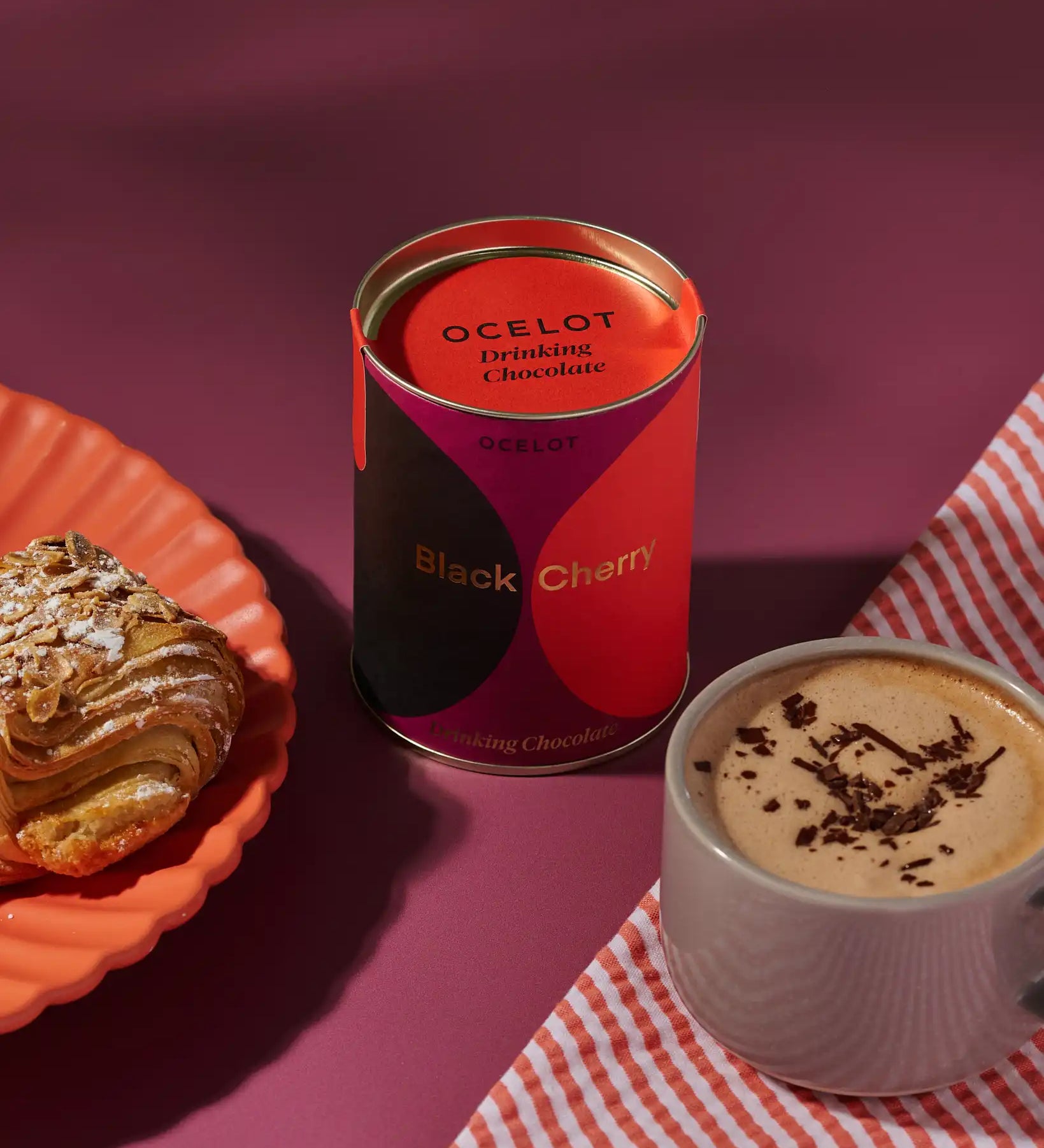 Ocelot Black Cherry Organic Drinking Chocolate in a tin placed next to a cup of hot chocolate and a pastry. 