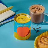 Ocelot Blood Orange Organic Drinking Chocolate in a 200g tin placed next to a cup of hot chocolate. and a cinnamon swirl. 