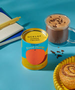 Ocelot Blood Orange Organic Drinking Chocolate in a 200g tin placed next to a cup of hot chocolate. and a cinnamon swirl. 