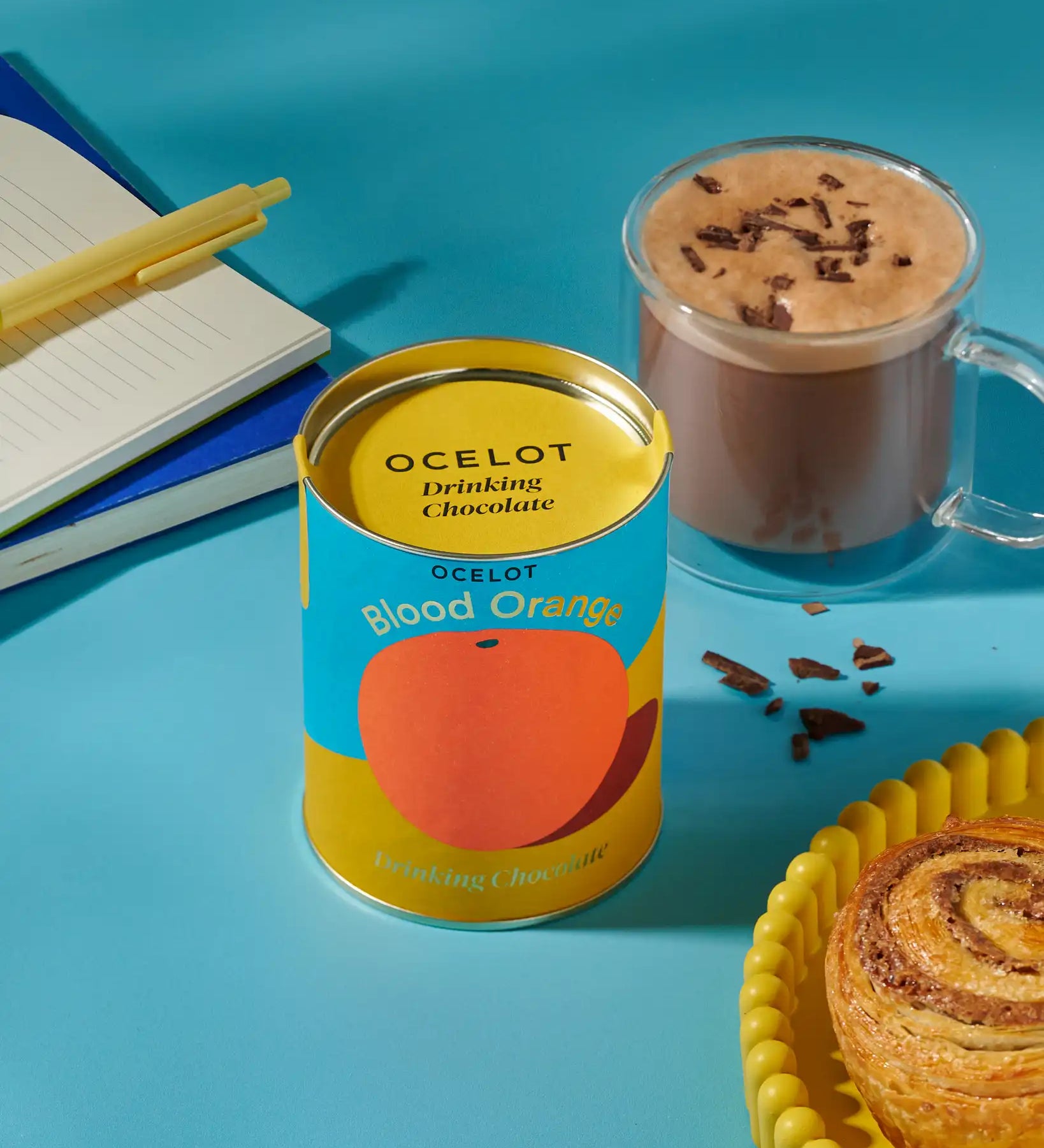 Ocelot Blood Orange Organic Drinking Chocolate in a 200g tin placed next to a cup of hot chocolate. and a cinnamon swirl. 