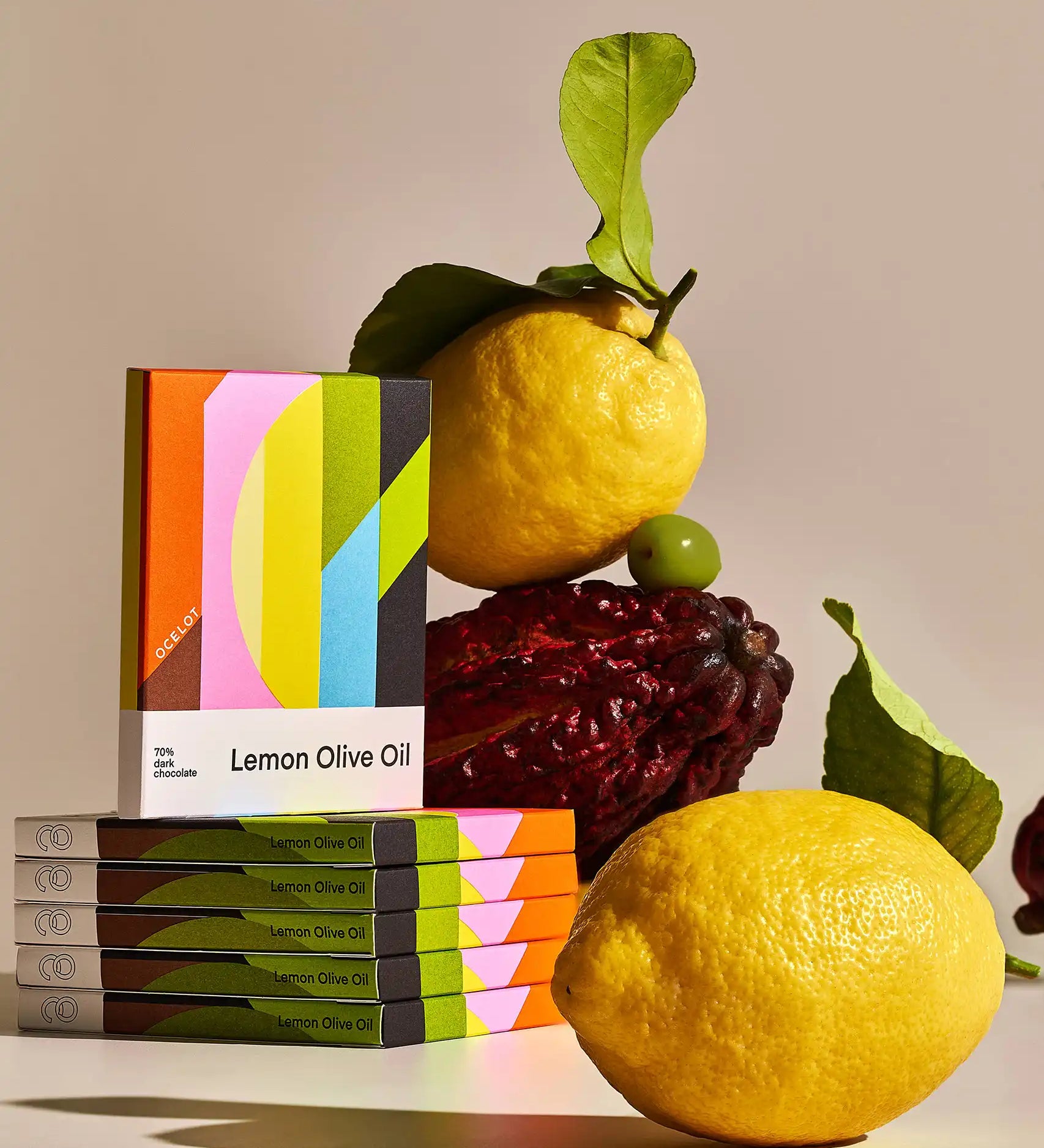 Bars of Ocelot Lemon Olive Oil Organic 70% Dark Chocolate in their boxes stacked up next to lemons, olives and a cacao pod. 