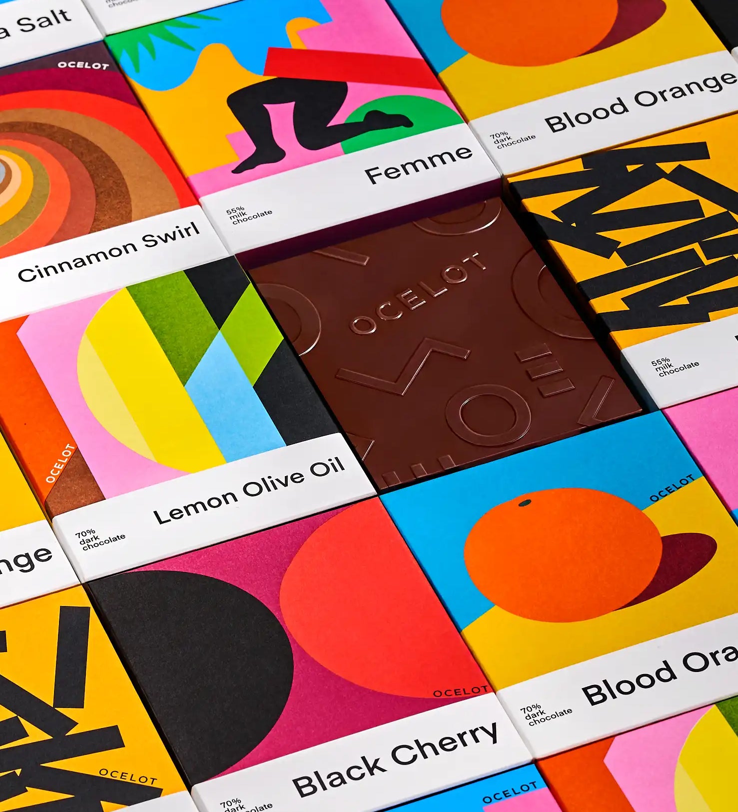 Various flavours of Ocelot chocolate bars in their modern, geometric style packaging boxes placed next to each other. Cinnamon swirl, femme, lemon olive oil, blood orange and black cherry varieties. One bar has been unwrapped.